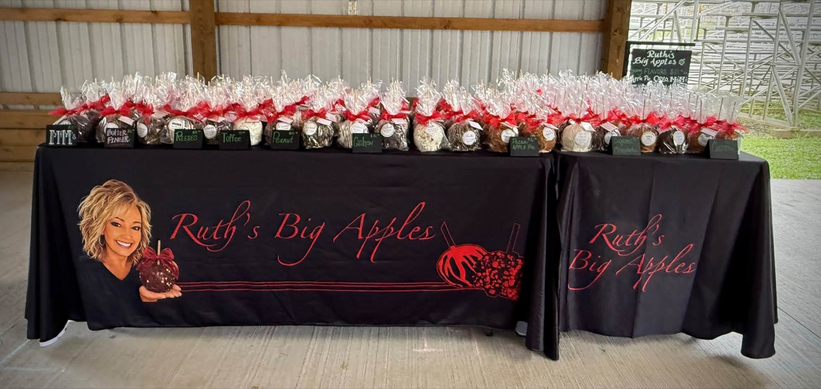 Two market tables covered in black cloths printed with Ruth's Big Apples, lined with bagged apples and chalk flavor signs