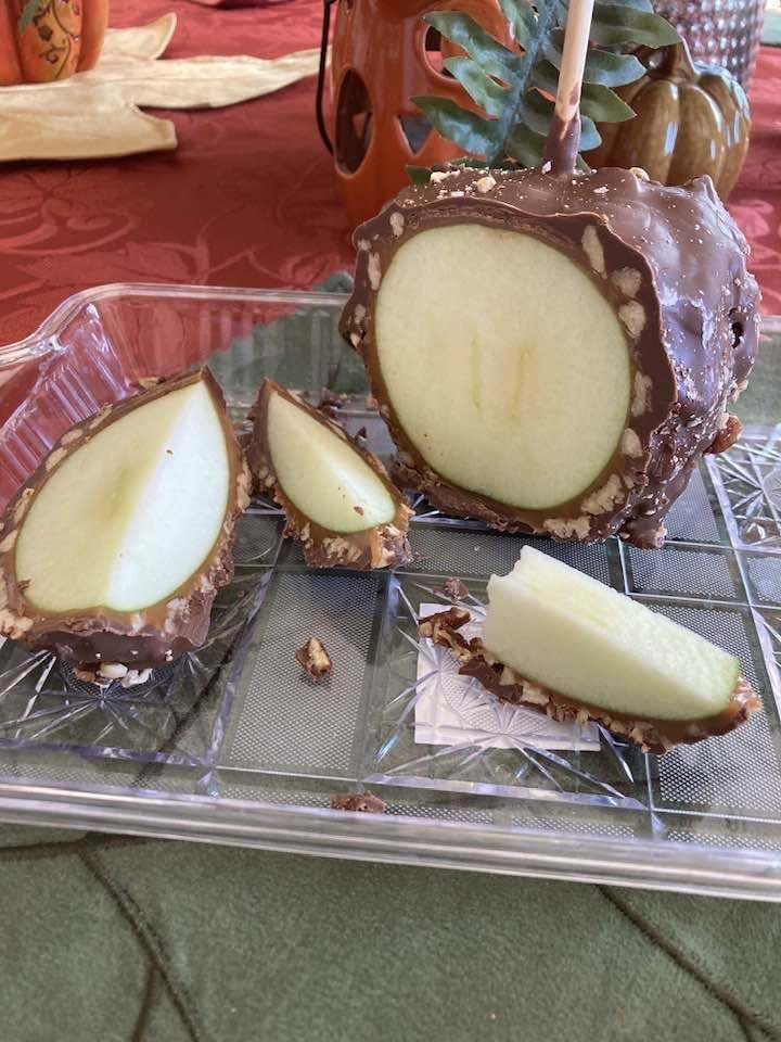 A nut-covered chocolate caramel apple sliced open on a glass tray, showing the caramel layer around a green apple