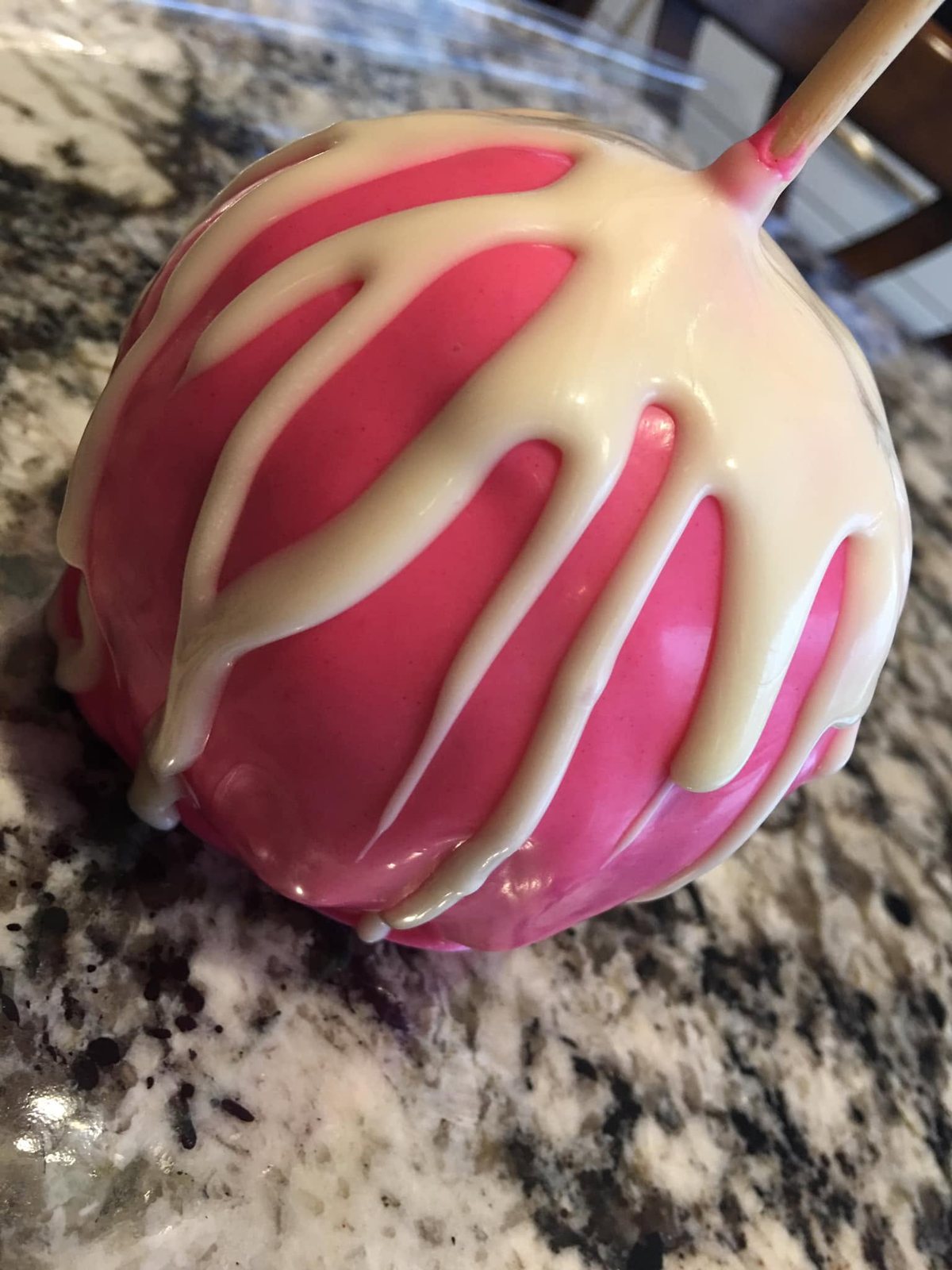 A pink-coated candy apple with a thick white drizzle, on a granite kitchen counter